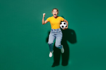 Naklejka na ściany i meble Full body young woman fan wear yellow t-shirt cheer up support football sport team hold in hand soccer ball watch tv live stream jump high do winner gesture scream isolated on dark green background.