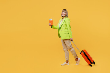 Full body elderly woman 50s years old in green jacket white t-shirt hold valise passport ticket isolated on plain yellow background. Tourist travel abroad in free time rest. Air flight trip concept.