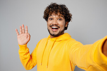 Close up young happy Indian man 20s he wearing casual yellow hoody doing selfie shot pov on mobile cell phone waving hand isolated on plain grey background studio portrait. People lifestyle portrait.