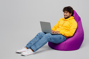 Full body young Indian IT man 20s he wearing casual yellow hoody sit in bag chair hold use working on laptop pc computer isolated on plain grey background studio portrait. People lifestyle portrait.