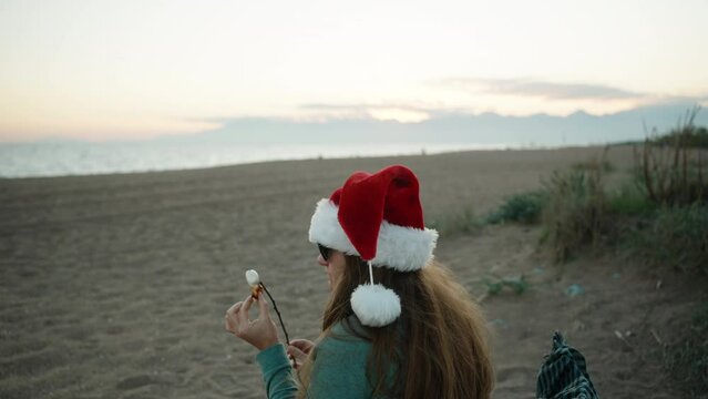 A girl on the beach in Santa's red hat is eating fried marshmallows, blowing on it.