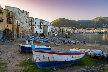 Fototapeta na wymiar Sunrise. Mediterranean coast. The old town of Cefalu. View of old boats, beaches with folded deckchairs. Mountains and the old town in the distance. Sicily. Italy.