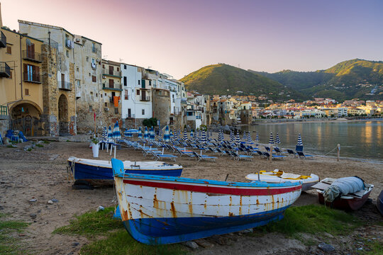 Sunrise. Mediterranean coast. The old town of Cefalu. View of old boats, beaches with folded deckchairs. Mountains and the old town in the distance. Sicily. Italy.