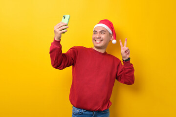 Fototapeta na wymiar Cheerful young Asian man in Santa hat taking selfie on smartphone, showing victory sign over yellow studio background. celebration Christmas holiday and New Year concept