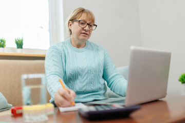 Happy mature businesswoman accountant in glasses and turquoise jumper sitting on the couch with laptop, taking notes, holding papers, contract studying or working remotely online from home