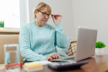 Happy mature businesswoman accountant in glasses and turquoise jumper sitting on the couch with laptop, taking notes, holding papers, contract studying or working remotely online from home