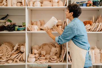 Mature woman taking clay jug from shelf while standing in workshop