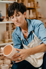 Mature white potter woman holding ceramic jug at the workshop indoors