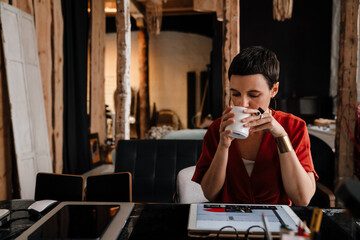 Young woman designer working on tablet and drinking coffee in cozy workshop