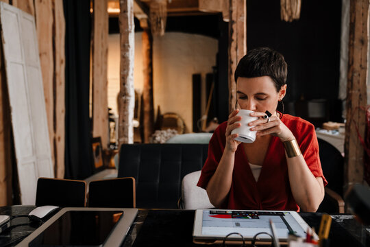 Young woman designer working on tablet and drinking coffee in cozy workshop