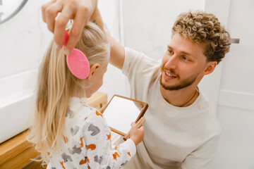 Young father helping his little daughter to comb her hair in bathroom