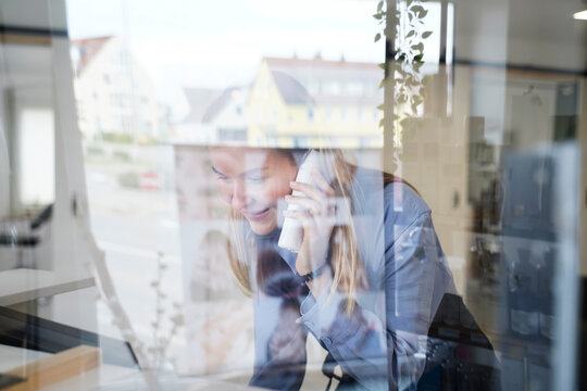 Smiling woman talking on phone seen through glass window in hair salon