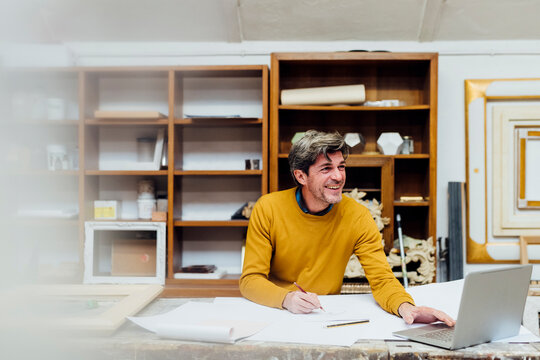 Happy mature craftsman with laptop and blueprint on workbench