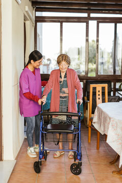 Smiling young caregiver helping senior woman to walk with mobility walker at home