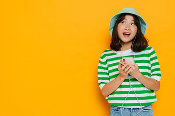 Young asian girl listening music with earphones using mobile phone isolated over yellow background