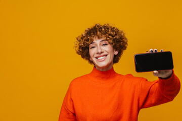 Happy middle-aged ginger woman showing black screen smartphone at camera while standing isolated over yellow wall