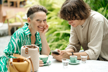 Concept for dating site or phone app. Man and woman in a cafe. The man looks at the screen of the phone, the woman at the camera and smiles.