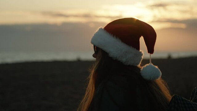 A girl in a red Santa hat looks at the sea. Christmas sunset on the background of mountains.