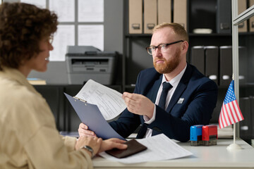 Portrait of bearded male consultant working in US embassy office and holding application form while talking to woman