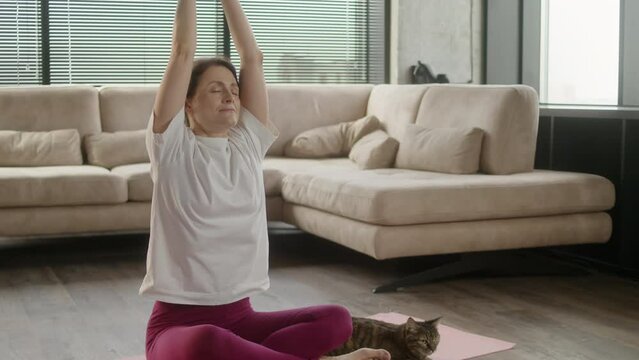 Middle age woman in sportswear practicing yoga at home with a cat at home. Female sit on mat in lotus position on red fitness mat in living room, healthcare and exercise mental therapy at home concept