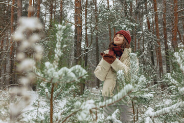 Smiling woman in winter outerwear enjoying snowfall in evergreen forest and looking up 
