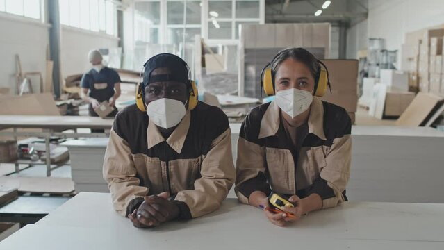 Portrait of multiethnic male and female workers in protective ear muffs and masks leaning on wooden plank and posing together for camera at furniture factory