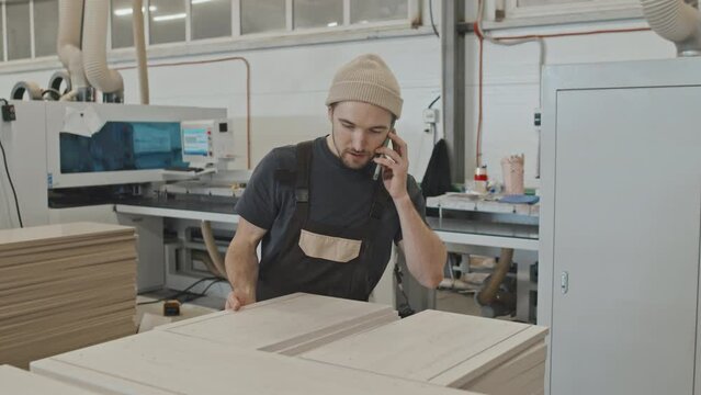 Man examining wooden planks and speaking on mobile phone while working in furniture factory