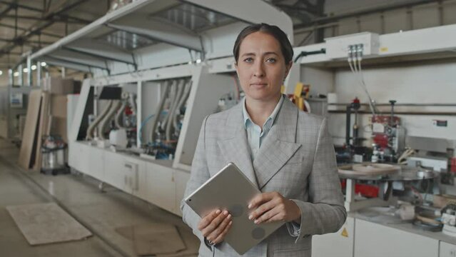 Medium waist up shot of female manager in formalwear holding digital tablet and posing for camera at work in furniture factory. Suitable for deepfake generation or face swap