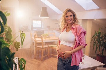 Portrait of pregnant woman standing at home holding hands on her belly