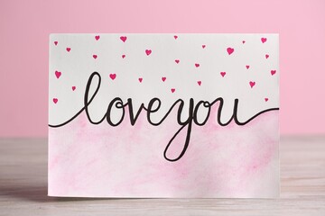 Card with phrase Love You and little drawn hearts on white wooden table against pink background