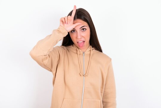 Funny Young caucasian woman wearing sweatshirt over white background makes loser gesture mocking at someone sticks out tongue making grimace face.
