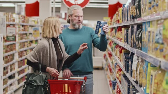 Mid adult Caucasian couple carrying shopping cart while walking along shelves with grocery products in supermarket choosing pasta