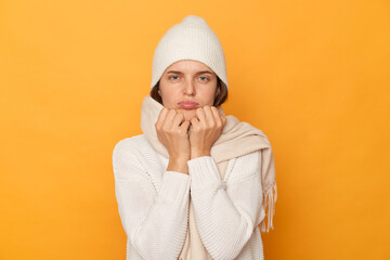 Horizontal shot of sad upset Caucasian woman wearing warm jumper, cap and scarf posing isolated over yellow background, standing with pout lips, being in bad mood.