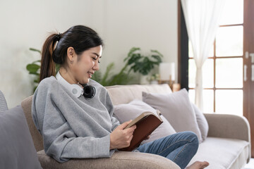 Young beautiful asian woman student sitting on sofa reading book at home.