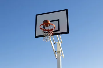 Gardinen Low angle shot of orange basketball flying into hoop outside on sunny day. Ball thrown into basket with white backboard against blue cloudless sky. Achievement, sports concept. © KAMPUS
