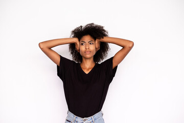 Portrait of frowning young woman covering ears with hands over white background. African American lady wearing black T-shirt and jeans protecting ears from noise. Stress and noise concept