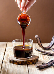 Healthy carob molasses and woman hand pouring liquid to the cup from glass jar, carob pods on wooden and brown background 