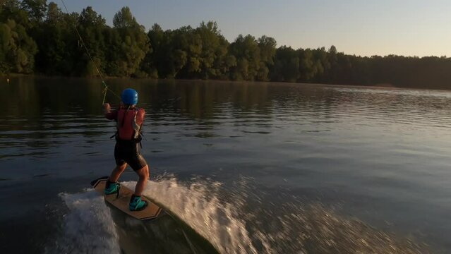 FPV drone flying following to extreme female wakeboard surfer wearing helmet riding on lake water at cable wake park on background of bright sunlight at summer sunny day. Tracking shot in slow motion.