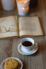 Plate with peanut butter cookies, cup of tea or coffee, open book, reading glasses and lit candles on the table. Selective focus.