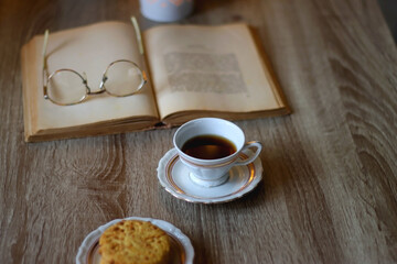 Plate with peanut butter cookies, cup of tea or coffee, open book, reading glasses and lit candles on the table. Selective focus.