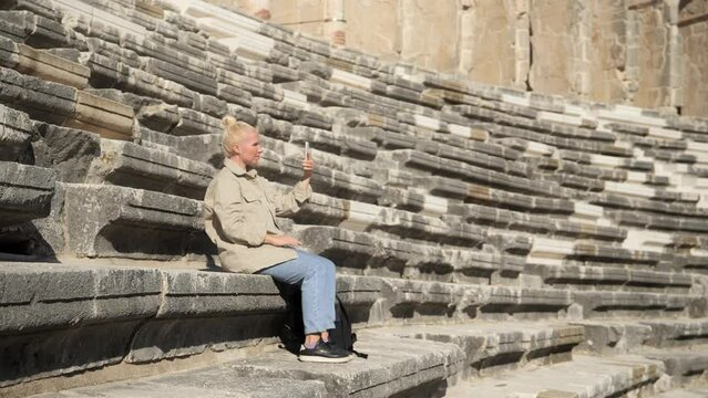 A young female student is sitting on the steps of an ancient amphitheater and taking pictures on her phone. Student of the Faculty of History. High quality 4k footage