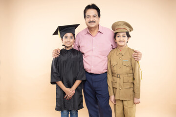 Happy senior Indian man standing with girl child wearing convocation costume and boy kid wearing...