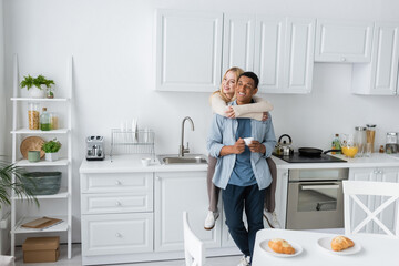 happy woman sitting on worktop in modern kitchen and embracing african american boyfriend with coffee cup