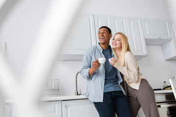 excited interracial couple with coffee cups looking way in kitchen on blurred foreground