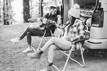 Fototapeta na wymiar Multiracial women friends having fun cheering with coffee camping outside camper van - Focus on african female hand holding cup - Black and white editing
