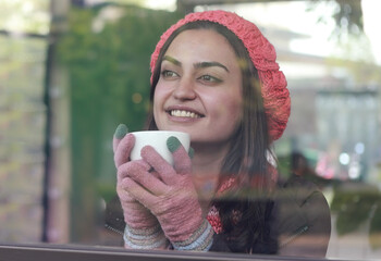 Looking through the glass, young woman in jacket sitting in cafe, smiling, looking at window. People lifestyle concept.