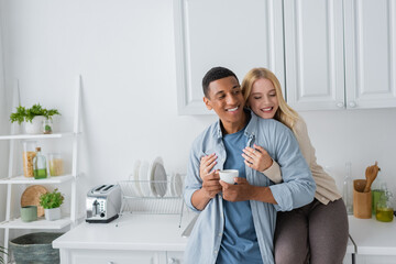 joyful blonde woman with closed eyes sitting on kitchen worktop and hugging african american boyfriend standing with coffee cup