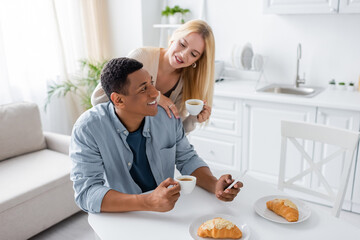 blonde woman holding coffee cup near african american man using smartphone during breakfast in...