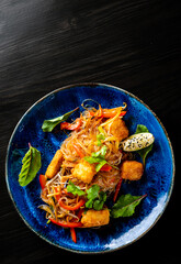 asian noodles with fried cheese tofu and vegetables in plate on black wooden table background