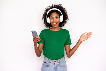 Portrait of happy young woman in headphones holding mobile phone over white background. African American lady wearing green T-shirt and jeans listening to phone player. Mobile technology concept